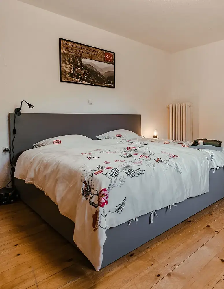 Schlafzimmer große Auszeit mit Blick auf graues Polsterbett mit floraler Tagesdecke und rustikalem Holzboden