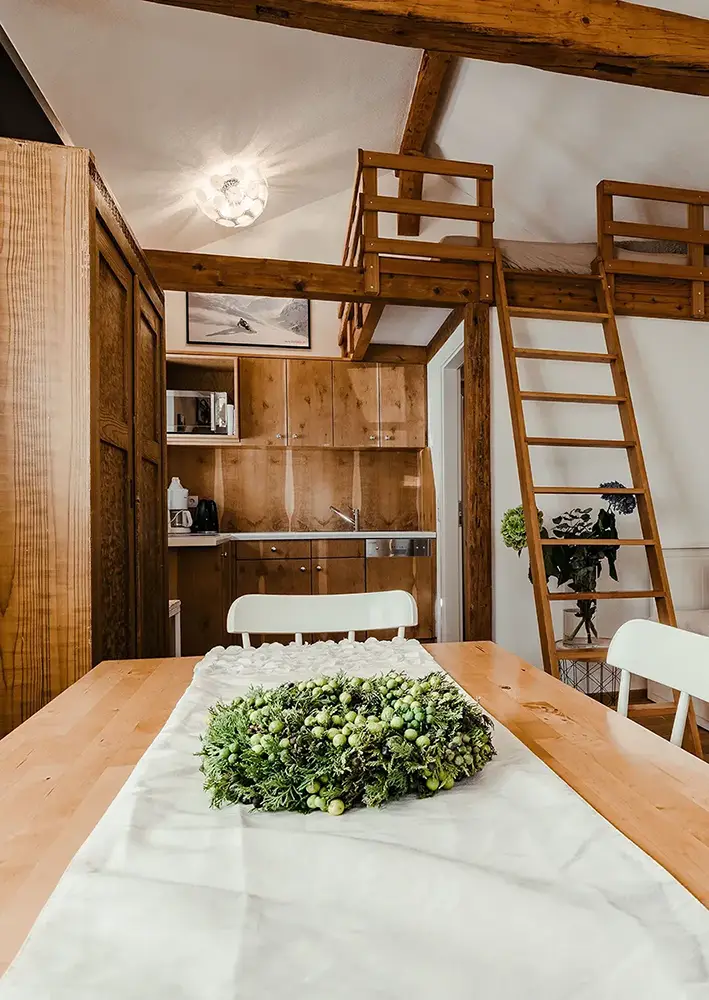 Gemütliches kleines Ferienhaus mit Holzinterieur, Blick von Esstisch mit Dekokranz auf kleine Küchenzeile und Leiter zum Schlafloft unter sichtbarem Holzbalken.